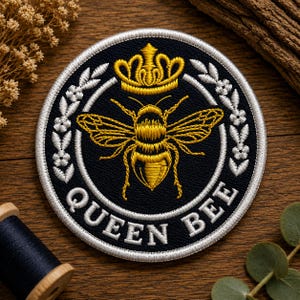 May include: A circular embroidered patch featuring a black background with a central golden bee topped by a golden crown. The design is framed by white floral wreaths and the text QUEEN BEE in white capital letters along the bottom edge.