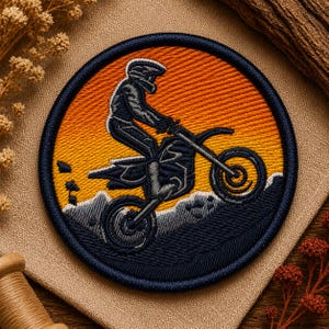 Pode incluir: Um emblema bordado circular com a silhueta cinzento-escura de uma pessoa numa mota de cross contra um pôr do sol laranja e amarelo. O design inclui uma cordilheira cinzento-escura na parte inferior e uma borda azul-marinho espessa.