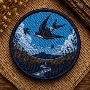 May include: A circular embroidered patch featuring a landscape scene with a dark blue sky, white clouds, and a black swallow in flight. Below, a dark mountain range sits above a winding light blue river, with dark brown hills and silhouettes of tall grass.