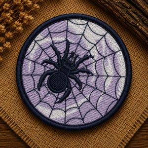 May include: A circular embroidered patch featuring a black spider centred on a light purple background with a white stitched spiderweb pattern. The patch has a dark navy blue border and is designed with a flat, round shape for application.