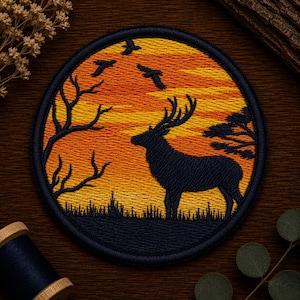 May include: A circular embroidered patch featuring a dark silhouette of a stag standing in a field against a vibrant orange and yellow sunset sky. The design includes a bare tree on the left, a pine tree on the right, and three birds flying overhead.