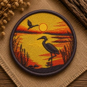 Puede incluir: Un parche bordado circular que muestra un paisaje al atardecer con una garza oscura en primer plano, un ave volando arriba y juncos altos a los lados. La escena utiliza hilos amarillos, naranjas y rojos vibrantes con un borde oscuro.