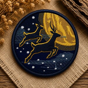 May include: A circular embroidered patch featuring two dark blue deer leaping across a night sky. A large, golden yellow moon sits in the background, while small white embroidered dots represent stars or snowflakes against the dark blue fabric.