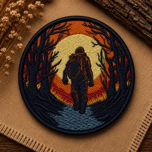 May include: A circular embroidered patch featuring a dark silhouette of a person with a backpack walking through a forest. The background displays a large yellow sun, orange sky, and dark blue trees. The ground is depicted with blue and yellow stitching.