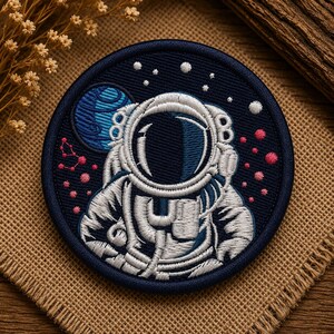 Może przedstawiać: Okrągła haftowana naszywka przedstawiająca białego astronautę w skafandrze kosmicznym na ciemnogranatowym tle. Wzór zawiera niebieską planetę, małe białe gwiazdy oraz skupiska drobnych różowych kropek przedstawiających odległe mgławice.