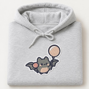 Puede incluir: Una sudadera con capucha gris claro que presenta un parche bordado de un murciélago pequeño, redondo y de color gris oscuro con el vientre de color tostado claro. El murciélago sostiene un globo rosa pálido y una piruleta roja y blanca.