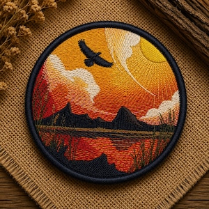May include: A circular embroidered patch featuring a landscape scene with a dark silhouette of a bird flying over a lake and mountains. The sky is filled with vibrant orange and yellow sunset colors, white clouds, and a bright yellow sun.