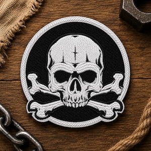May include: A circular embroidered patch featuring a white skull and crossbones design on a black background. The patch is bordered by a white rope-style edge. The skull has a small cross symbol on the forehead and detailed shading in white thread.