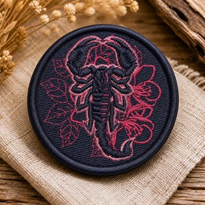 May include: A circular embroidered patch featuring a dark navy blue scorpion centered against a background of thin, magenta-colored floral and leaf outlines. The patch has a thick, dark navy blue stitched border and a dense, textured fabric base.