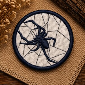 May include: A circular embroidered patch featuring a dark navy blue spider centered on a cream-colored background with a geometric spider web pattern. The patch has a thick, dark navy blue stitched border that frames the entire circular design.