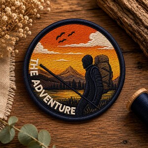 Puede incluir: Un parche bordado circular que muestra a un excursionista con una mochila grande y un bastón en un paisaje montañoso al atardecer. El cielo es naranja y amarillo con nubes blancas y pájaros. El texto THE ADVENTURE está bordado en blanco.