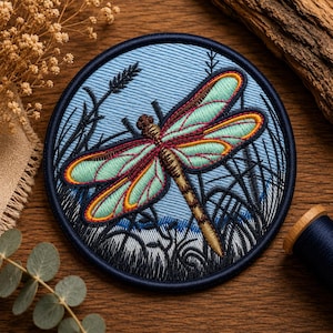 May include: A circular embroidered patch featuring a dragonfly with light green wings and a brown body. The background of the patch depicts a light blue sky with dark blue silhouettes of tall grasses and reeds, all enclosed by a dark blue border.
