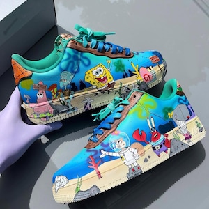 May include: A pair of low-top sneakers featuring hand-painted illustrations of Spongebob Squarepants characters including Patrick, Squidward, Mr. Krabs, Sandy, and Pearl. The shoes have a blue gradient background, teal laces, and a beige rubber sole.