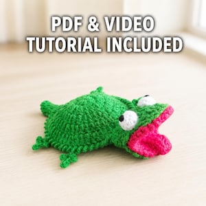 May include: A green crocheted frog toy with a bright pink tongue and white eyes with black pupils. The text PDF & VIDEO TUTORIAL INCLUDED is displayed above the frog. The frog has a textured yarn surface and four small limbs.