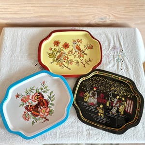 May include: Three rectangular metal trays with scalloped edges. One tray is yellow with birds and berries, one is white with a butterfly and flowers, and one is black with a detailed scene of figures in traditional clothing and trees.