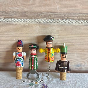 Peut inclure: Quatre figurines en bois peintes à la main servant d'outils de bar. De gauche à droite : un bouchon de bouteille féminin, un décapsuleur masculin, un tire-bouchon masculin aux bras jaunes et un bouchon de bouteille masculin au chapeau vert.