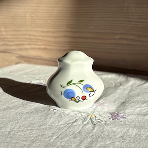 Op de afbeelding: Een witte keramische zout- of peperstrooier met een ronde, bolvormige vorm. De voorzijde is voorzien van een handgeschilderd bloemmotief bestaande uit twee blauwe cirkelvormige bloemblaadjes, een klein geel centrum, een rode stip en groene bladeren met zwarte steeltjes.