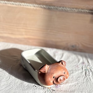 May include: A rectangular ceramic tray featuring a sculpted pig head at one end. The tray has a cream-colored interior and exterior, while the pig head is finished in a warm, reddish-brown glaze with detailed ears, eyes, and a snout.