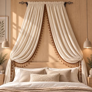 May include: A pair of off-white curtains draped over a bed headboard, featuring a brown pom-pom trim along the inner edges. The curtains are suspended from a black rod and hang down to the sides of the bed, which is styled with several pillows.