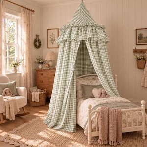 May include: A sage green and white gingham fabric bed canopy featuring a ruffled top edge, suspended over a twin-sized bed frame. The canopy drapes down to the floor, creating a tent-like structure around the bed, which is dressed in patterned bedding.
