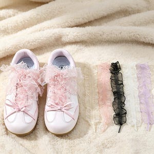 May include: A pair of white sneakers with light pink accents and pink ruffled lace shoelaces tied in bows, accompanied by five additional sets of ruffled lace shoelaces in cream, light pink, black, white, and lavender colors.