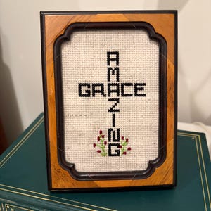 May include: A framed cross-stitch embroidery on light-coloured fabric featuring the black text "AMAZING GRACE" arranged in a cross shape. Small red and green floral accents are stitched at the base. The piece is enclosed in a dark wood frame.