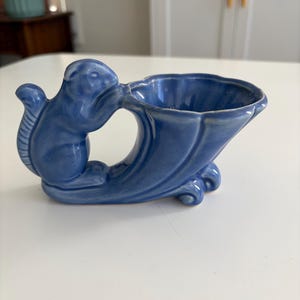 Może przedstawiać: Błyszcząca ceramiczna doniczka w kształcie rogu obfitości z niebieską glazurą. Naczynie posiada wyrzeźbioną figurkę wiewiórki przymocowaną z boku, rozszerzający się, falowany otwór oraz zakrzywioną podstawę, która zapewnia stabilność przedmiotu.