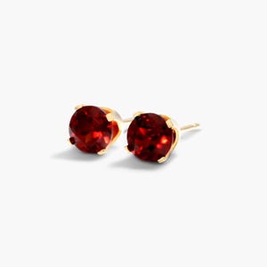 Puede incluir: Un par de pendientes de botón con piedras preciosas redondas de color rojo intenso facetadas, engastadas en garras de metal dorado. Cada pendiente tiene un cierre de poste, destacando el color vibrante y el brillo de las piedras sobre el metal.