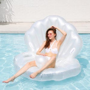 May include: A large, iridescent white inflatable pool float shaped like an open seashell. The float features a high, curved backrest and a wide, rounded base. A matching, spherical white inflatable ball is positioned in the center of the seat.