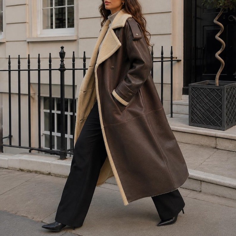 May include: A long, dark brown coat with a cream-colored shearling lining, wide lapels, and shoulder epaulets. The coat is worn over a light gray turtleneck sweater and black wide-leg trousers, paired with black pointed-toe heeled boots.