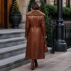 May include: A long, brown leather coat featuring a large, plush fur collar and matching fur cuffs. The coat has a structured, tailored fit with a defined waistline, a central back vent, and visible seam detailing. A brown leather handbag is held at the side.