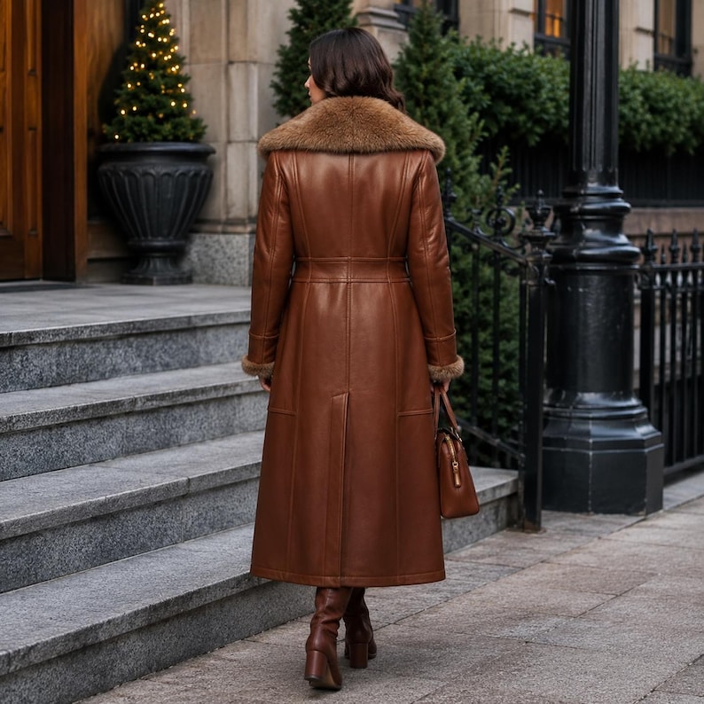 May include: A long, brown leather coat featuring a large, plush fur collar and matching fur cuffs. The coat has a structured, tailored fit with a defined waistline, a central back vent, and visible seam detailing. A brown leather handbag is held at the side.