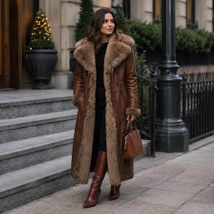 May include: A long brown leather coat featuring a thick, plush tan faux fur lining, a wide lapel collar, and long sleeves with fur cuffs. The coat is worn over a black top and black trousers, paired with brown leather knee-high boots and a brown handbag.