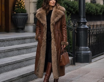May include: A long brown leather coat featuring a thick, plush tan faux fur lining, a wide lapel collar, and long sleeves with fur cuffs. The coat is worn over a black top and black trousers, paired with brown leather knee-high boots and a brown handbag.
