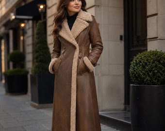 May include: A long brown coat featuring a tan faux shearling lining along the wide lapels, cuffs, and hem. The coat has a smooth exterior finish, long sleeves, and a structured silhouette that extends to the lower calf, worn over a black top.