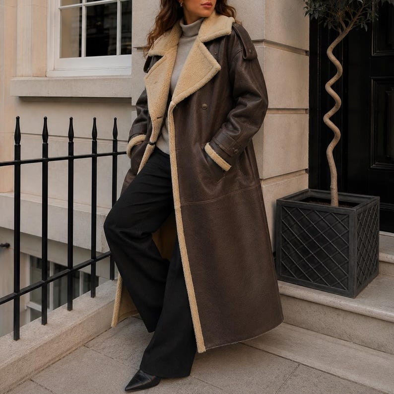 May include: A long dark brown leather coat featuring a wide cream-colored shearling collar and lapels, long sleeves with cuff straps, and a double-breasted button closure. The coat is worn over a light gray turtleneck sweater and black trousers.