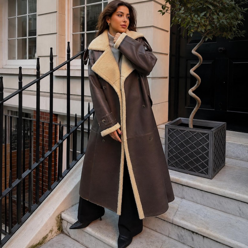 May include: A long, dark brown coat featuring a wide, cream-colored faux shearling lapel and collar. The coat has a double-breasted design with long sleeves, buttoned cuffs, and a smooth finish, worn over black trousers and black pointed-toe boots.