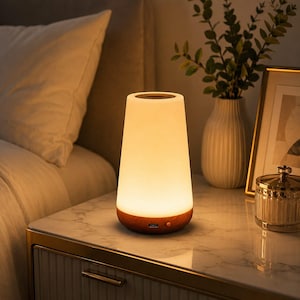 Puede incluir: Una lámpara de mesa cónica con una pantalla blanca translúcida y lisa que emite un brillo cálido y suave. La base está construida con un material de grano de madera oscuro y cuenta con un pequeño botón circular y un puerto de carga frontal.