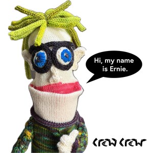 May include: A handmade knit sock puppet named Ernie with cream-colored skin, bright blue eyes, black circular glasses, and spiky lime green hair. The puppet wears a multi-colored textured sweater and features a speech bubble saying, Hi, my name is Ernie.