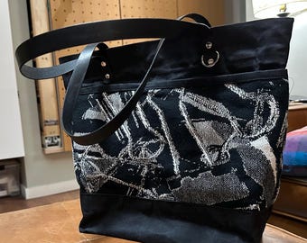 May include: A black tote bag featuring a textured, woven panel with a white abstract graphic pattern. The bag includes solid black fabric sections at the top and bottom, black leather-like straps attached with silver-toned metal rivets, and a D-ring.