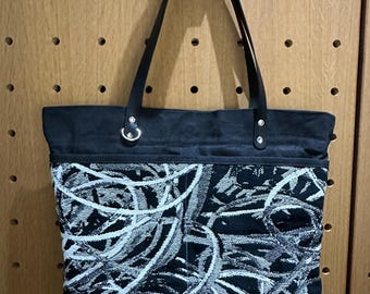 May include: A black tote bag featuring a central panel with a white and black abstract circular pattern. The bag has solid black fabric at the top and bottom, black shoulder straps attached with silver-toned metal rivets, and a silver D-ring.