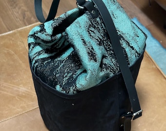 May include: A cylindrical bucket bag featuring a solid black base and a top section made of a woven fabric with a black and teal abstract pattern. The bag includes black straps attached with silver-toned metal hardware and a drawstring closure.