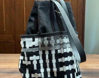 May include: A black bucket bag featuring a white and black geometric woven pattern on the lower exterior. The bag has a solid black upper section with a drawstring closure and a single black leather strap attached with metal hardware rings.