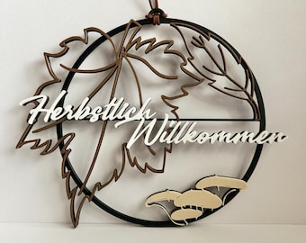 May include: A circular wall hanging featuring a black frame with a brown maple leaf cutout, light-colored mushroom shapes at the bottom, and the white text "Herbstlich Willkommen" across the center, suspended by a brown leather cord.