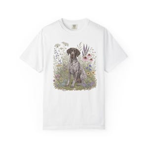 Op de afbeelding: Een wit t-shirt met korte mouwen en een centrale afbeelding van een bruin met wit gevlekte Duitse staande hond die tussen een kleurrijk assortiment wilde bloemen zit, waaronder paarse, roze en gele bloesems met groen blad.
