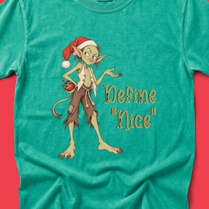 May include: A bright green t-shirt featuring a graphic of a green creature wearing a red Santa hat and brown tattered pants. The creature holds a red ornament. The text "Define "Nice"" is printed in black with a gold outline to the right.