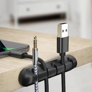Peut inclure: Un organisateur de câbles en silicone noir doté de cinq fentes fixé au bord d'un bureau. L'organisateur maintient un câble auxiliaire tressé argenté et blanc avec le texte Essager imprimé dessus et un câble USB noir avec un connecteur métallique.
