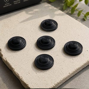 May include: Four circular black shock-absorbing pads with a tiered, stepped design. The packaging box features icons and text including SHOCK ABSORPTION, PROTECTS YOUR CAR, DURABLE MATERIAL, and EASY INSTALLATION, highlighting the product features.