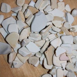 May include: A pile of white and cream-colored ceramic tiles, some with a slightly rough texture, on a light wood surface.