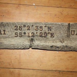 Könnte beinhalten: Ein verwittertes Holzschild mit dem Text "DUBAI 25° 2' 35" N 55° 12' 29" E UAE" eingebrannt in die Oberfläche.
